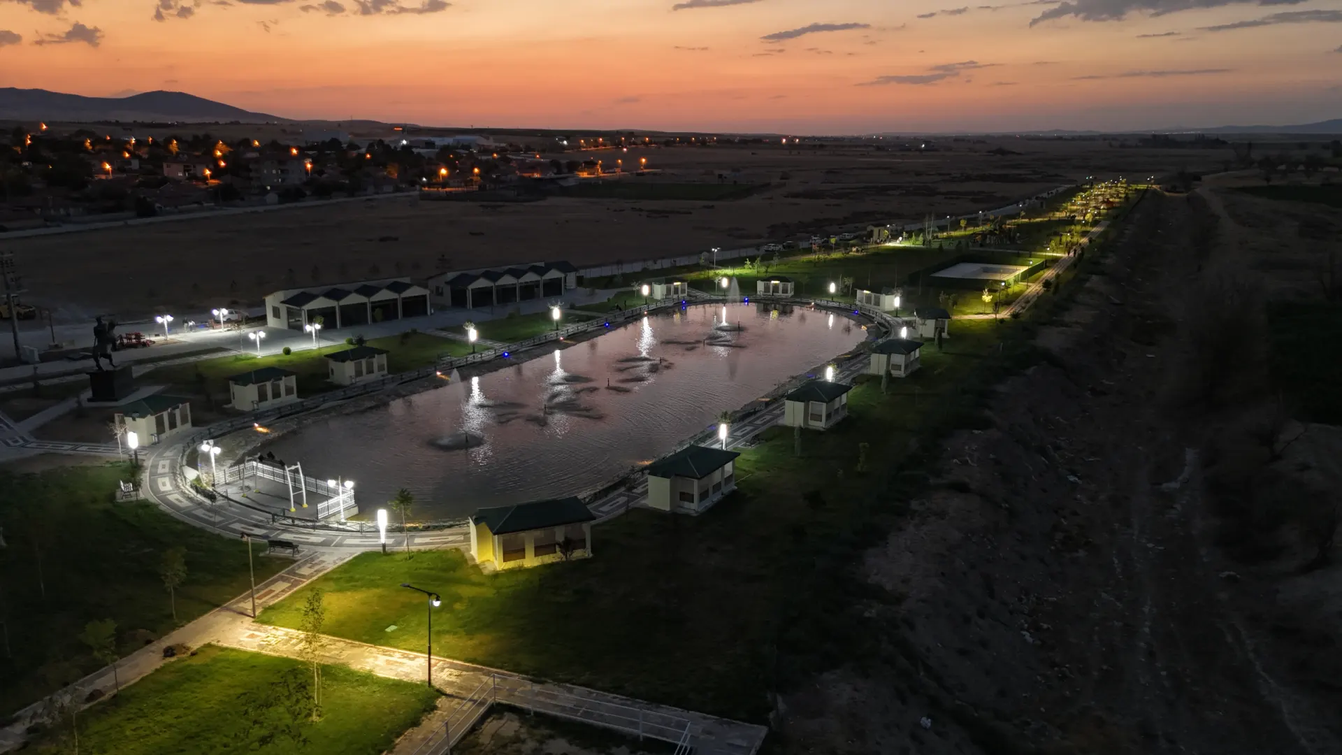 Göletli Park’ın akşam saatlerinde havadan görünümü: aydınlatılmış gölet, çevresindeki yürüyüş yolları ve uzakta kasabanın ışıkları